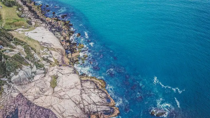 Costão rochoso e mar azul-turquesa vistos de cima, com a espuma das ondas nas pedras