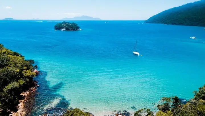 Enseada de água azul com um veleiro, ilha e costão coberto de mata
