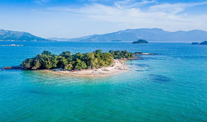 Ilha coberta de mata com faixa de areia branca, mar azul e montanhas ao fundo