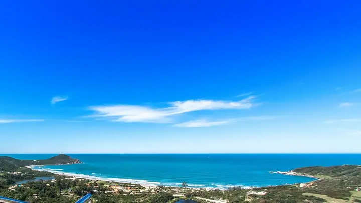 Panorama aéreo de praia em curva com morros, vegetação e céu azul com poucas nuvens