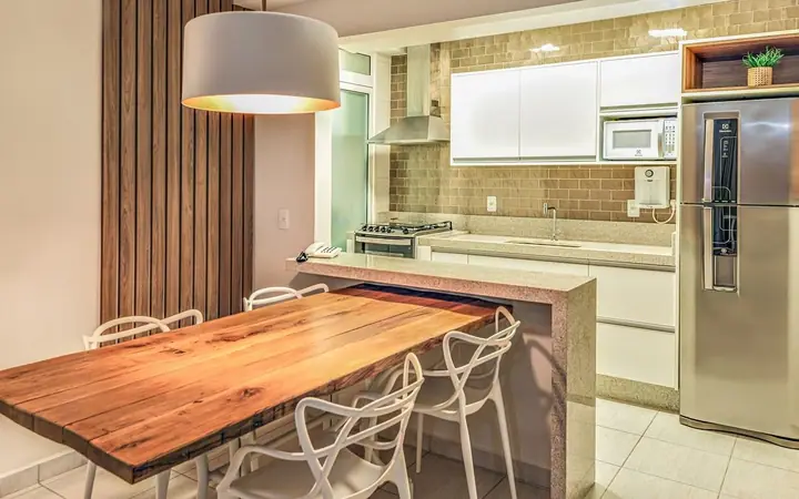 Cozinha de apartamento com mesa de madeira maciça, cadeiras brancas vazadas, pendente branco e armários claros