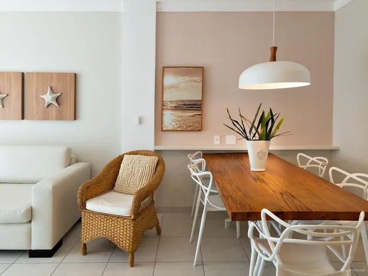 Mesa de jantar de madeira com cadeiras brancas, poltrona de fibra, pendente branco e quadro de praia