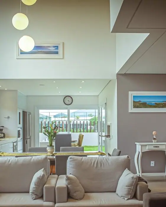 Living de casa contemporânea com pé-direito duplo, sofás claros, pendentes de vidro e cozinha integrada voltada para o jardim