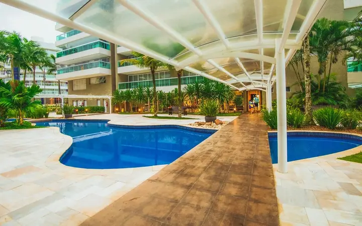 Área de lazer de condomínio com piscina de formas orgânicas, deck de madeira, pergolado de vidro e jardim tropical