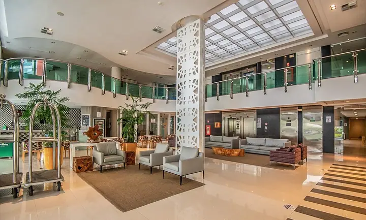 Lobby de hotel com claraboia, coluna vazada branca, mezanino com guarda-corpo de vidro, poltronas e carrinho de bagagem