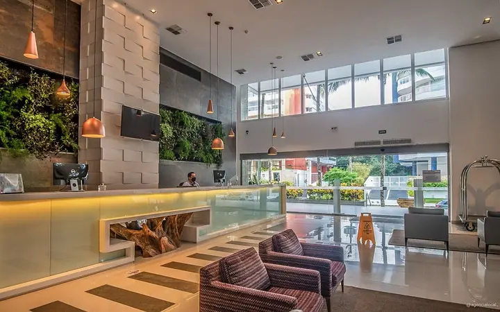 Recepção de hotel com pé-direito alto, jardim vertical, balcão iluminado e poltronas listradas