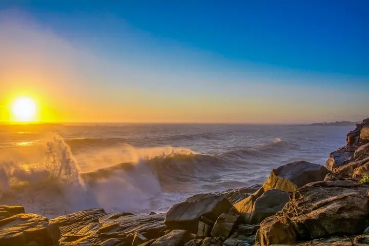 Onda quebrando nas pedras do costão contra o sol baixo, com o céu azul
