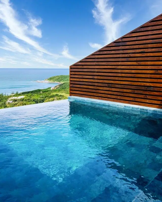 Piscina de borda infinita com parede de madeira ripada, o mar azul e a vegetação de restinga ao fundo