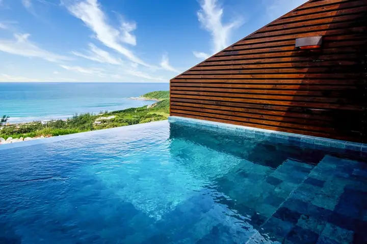 Piscina de borda infinita com parede de madeira ripada, o mar azul e a vegetação de restinga ao fundo
