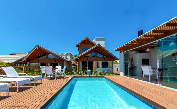 Piscina retangular com deck de madeira e espreguiçadeiras diante de chalés de madeira, sob céu azul