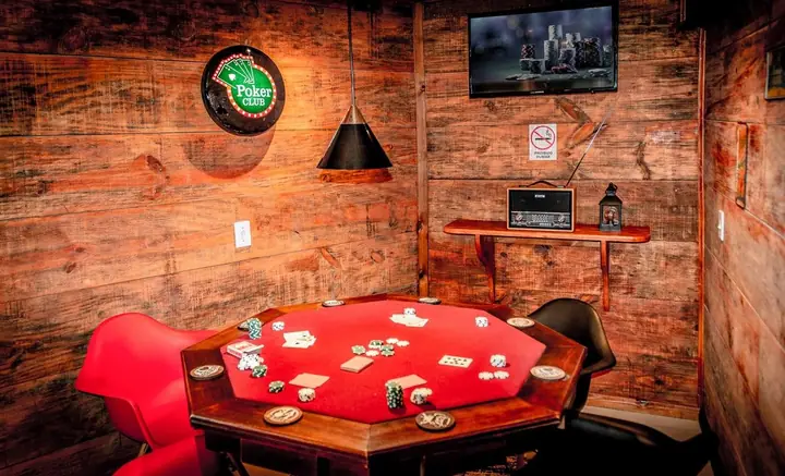 Sala de jogos com mesa de pôquer de feltro vermelho, cadeiras vermelhas, luminária pendente e paredes de madeira rústica