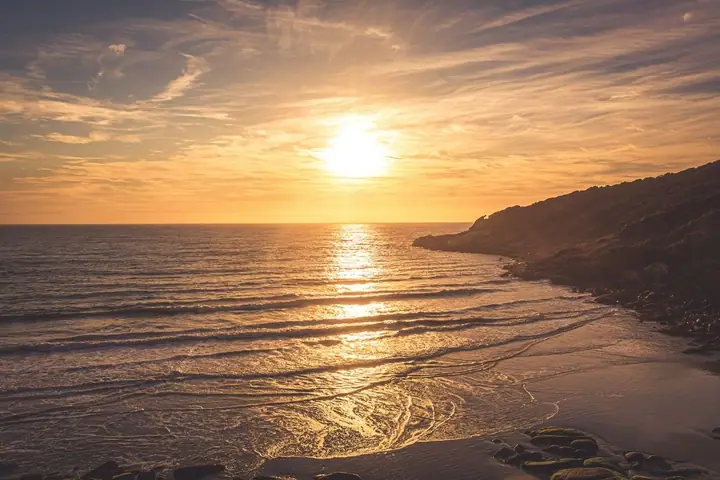 Sol baixo sobre o mar, com o costão escuro à direita e reflexos dourados na areia molhada