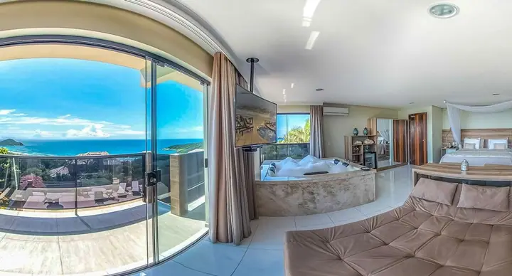 Suíte com banheira de hidromassagem junto à porta de vidro, com vista para o mar, as ilhas e os morros