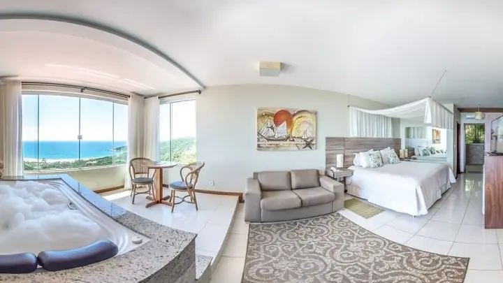 Suíte de pousada com banheira de hidromassagem junto à janela panorâmica com vista para o mar, sala de estar e cama com dossel